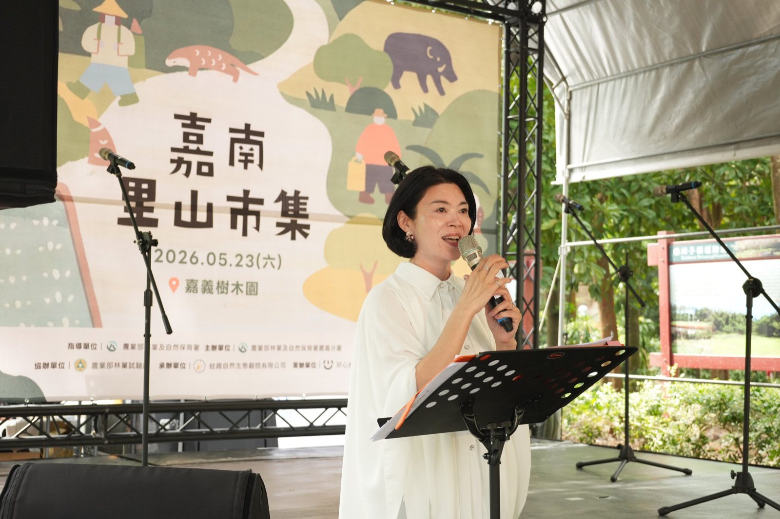 115年度諸羅里山暨生態農業市集開幕典禮，主持人於「嘉南里山市集」主視覺舞台背板前致詞