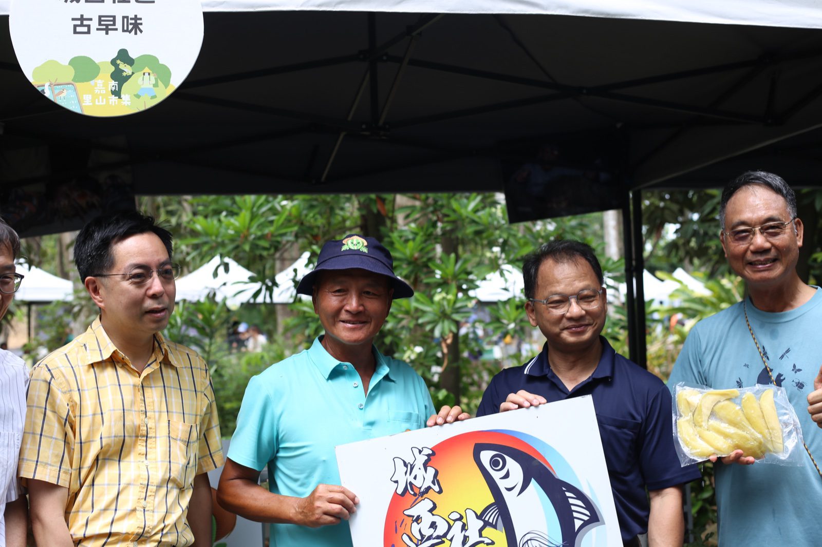 長官貴賓於市集攤位前與攤商合影，攤位圓牌印有嘉南里山市集主視覺