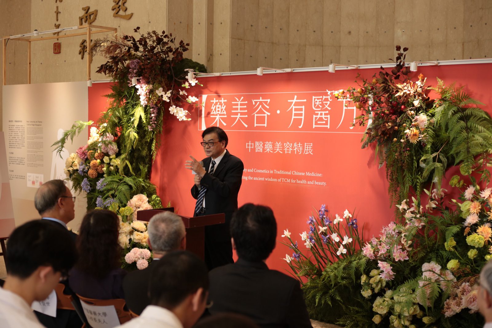 立夫中醫藥博物館「藥美容‧有醫方」特展開幕致詞,講者於花藝背板前致詞